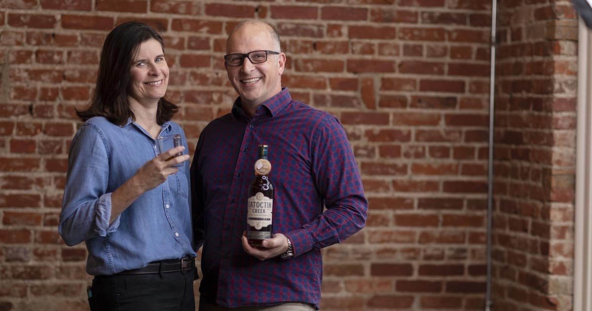 Becky and Scott Harris of Catoctin Creek Distilling, woman in a blue shirt and man in a red plaid shirt in front of a brick wall, woman is holding a glass of whiskey and man is holding a bottle that reads Catoctin Creek on the label