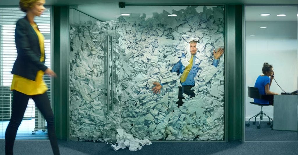 Time to go paperless: a man is surrounded by paper in a glass office